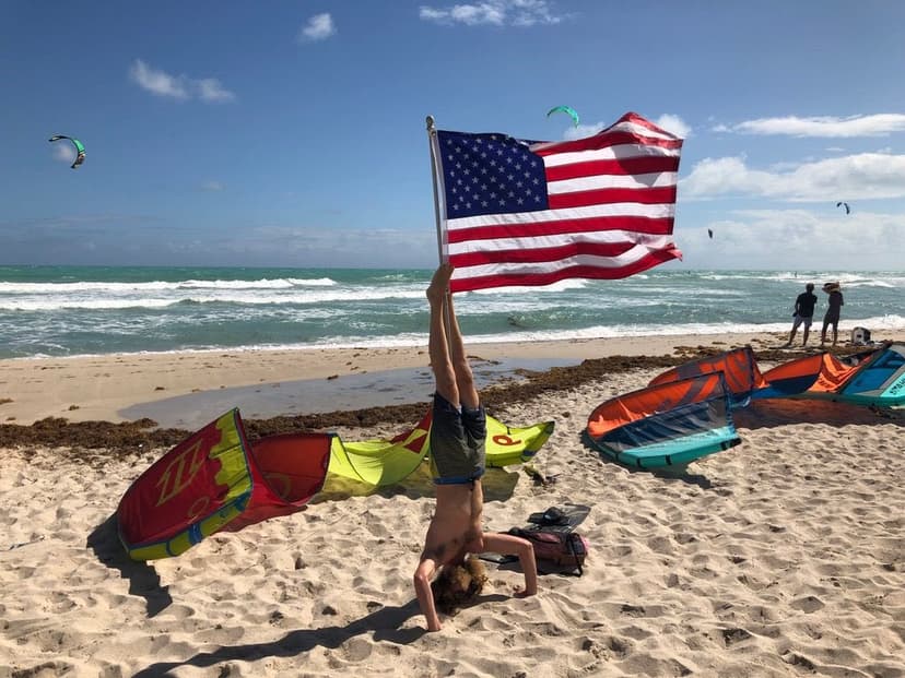 Glen Bradford doing a handstand on the beach holding an American flag with kites in the sky